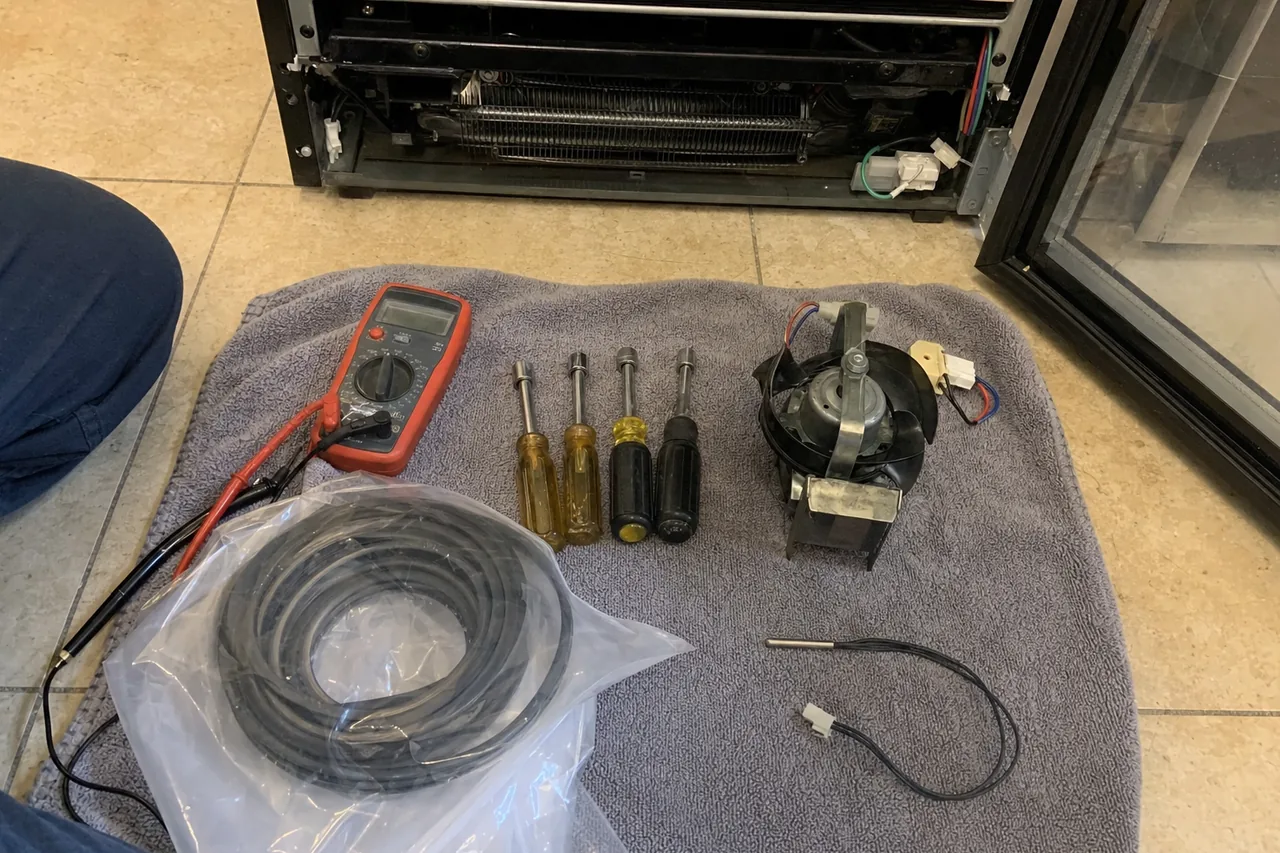 Multimeter, four nut drivers, an evaporator fan motor, a bagged door gasket and a temperature sensor laid out on a towel
