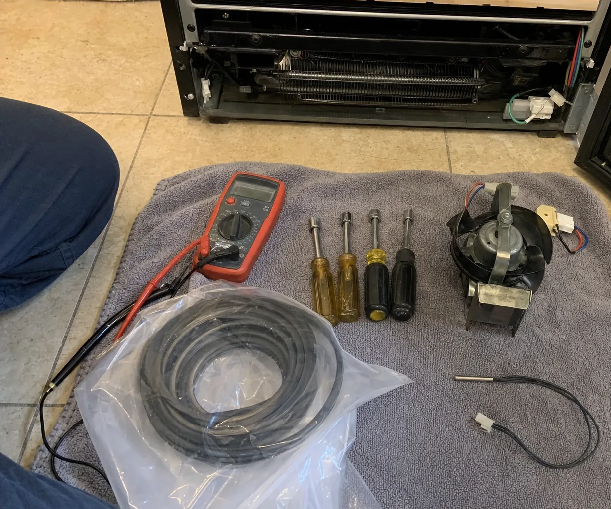 A digital multimeter with its red and black leads, and four nut drivers, ready on a towel beside an opened appliance