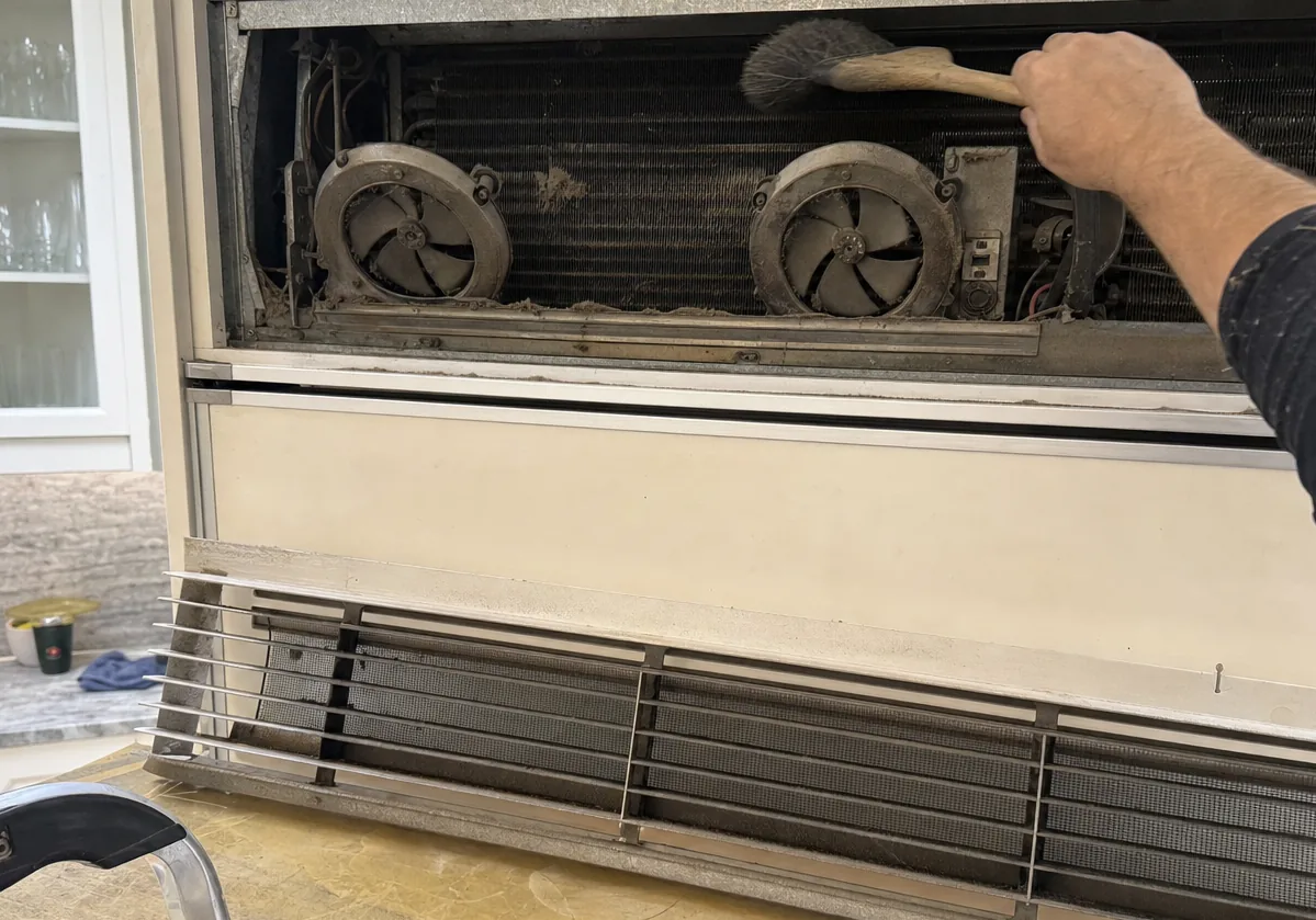 Close view of a lint-loaded condenser coil and its two fans, mid-brush, behind a built-in refrigerator grille