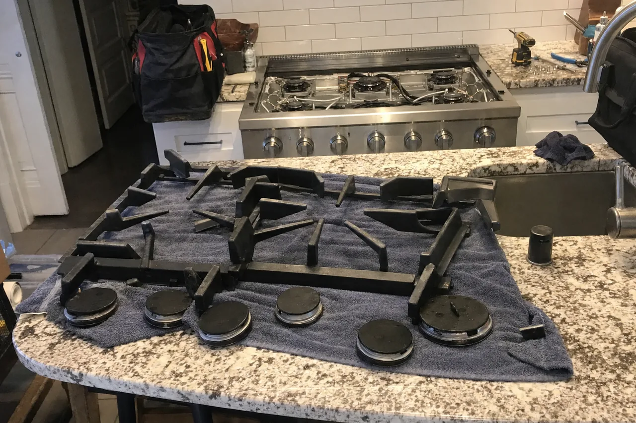 Cast-iron grates and burner caps laid out on a towel on a granite island, with a stripped stainless rangetop behind